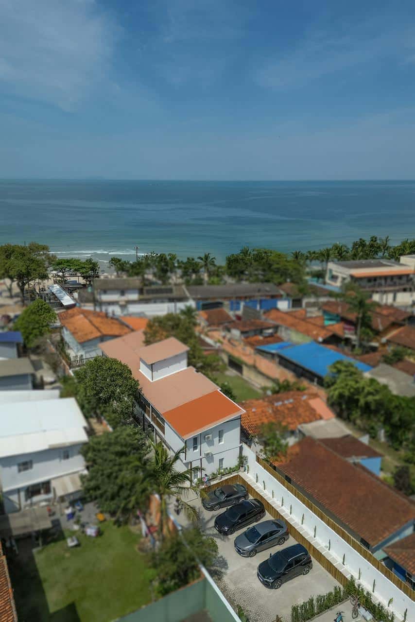 Vista aérea do bairro: o telhado de duas águas do hotel e o pátio de estacionamento em primeiro plano, os telhados de Maresias em volta, a faixa de areia e o mar aberto ocupando o fundo do quadro