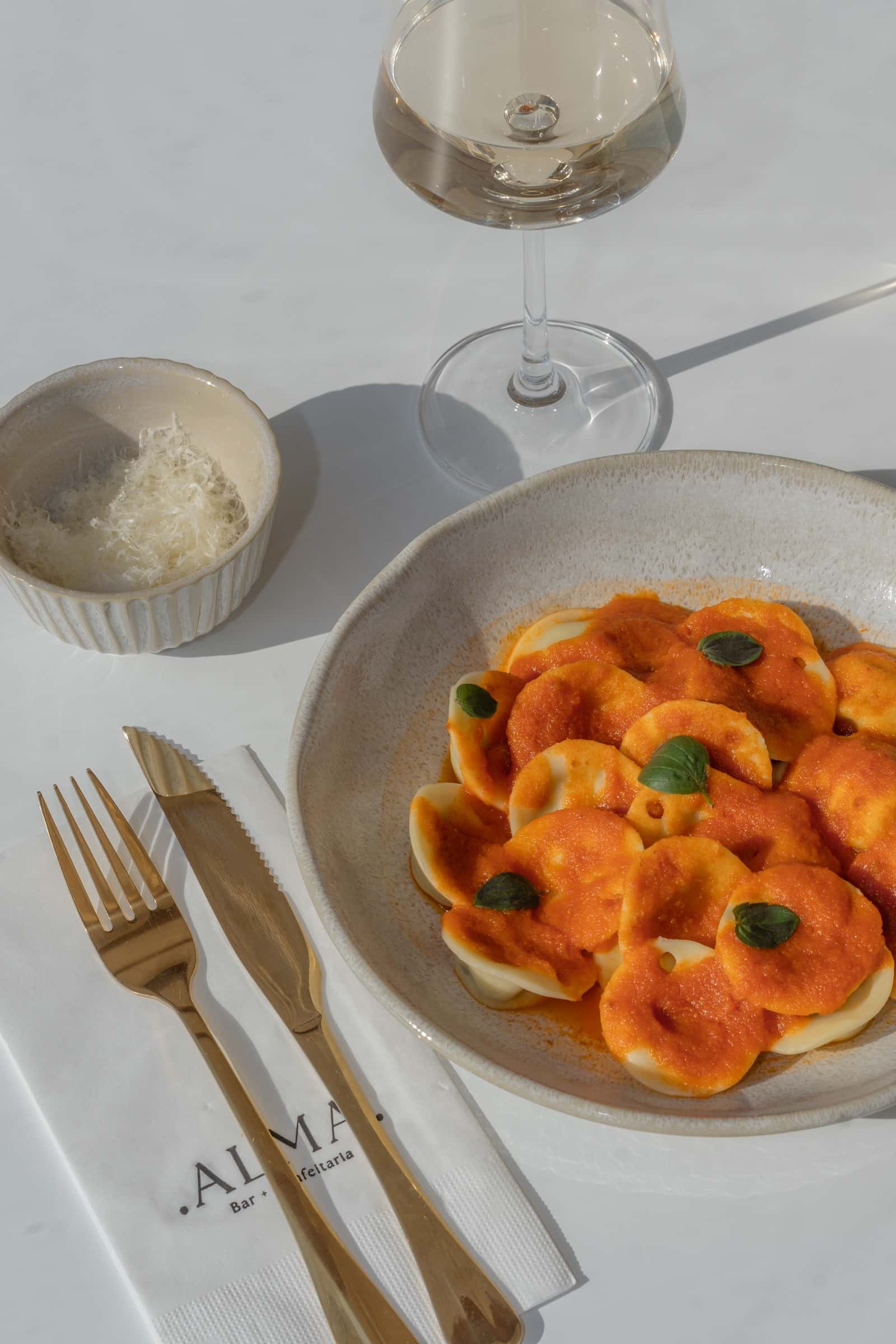 Massa recheada ao molho de tomate em prato de cerâmica, com queijo ralado, taça de vinho e guardanapo com o logotipo ALMA