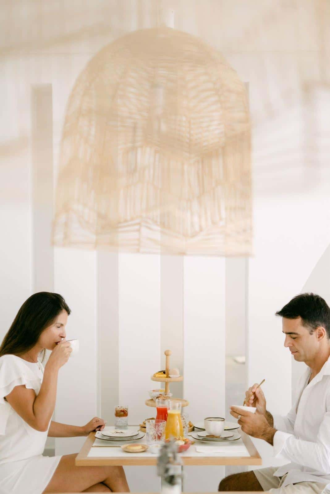 Casal à mesa do café da manhã no salão branco: ela toma café na xícara, ele se serve de uma tigela, com a torre de três andares, os sucos e a louça entre os dois e um grande pendente de fibra trançada por cima