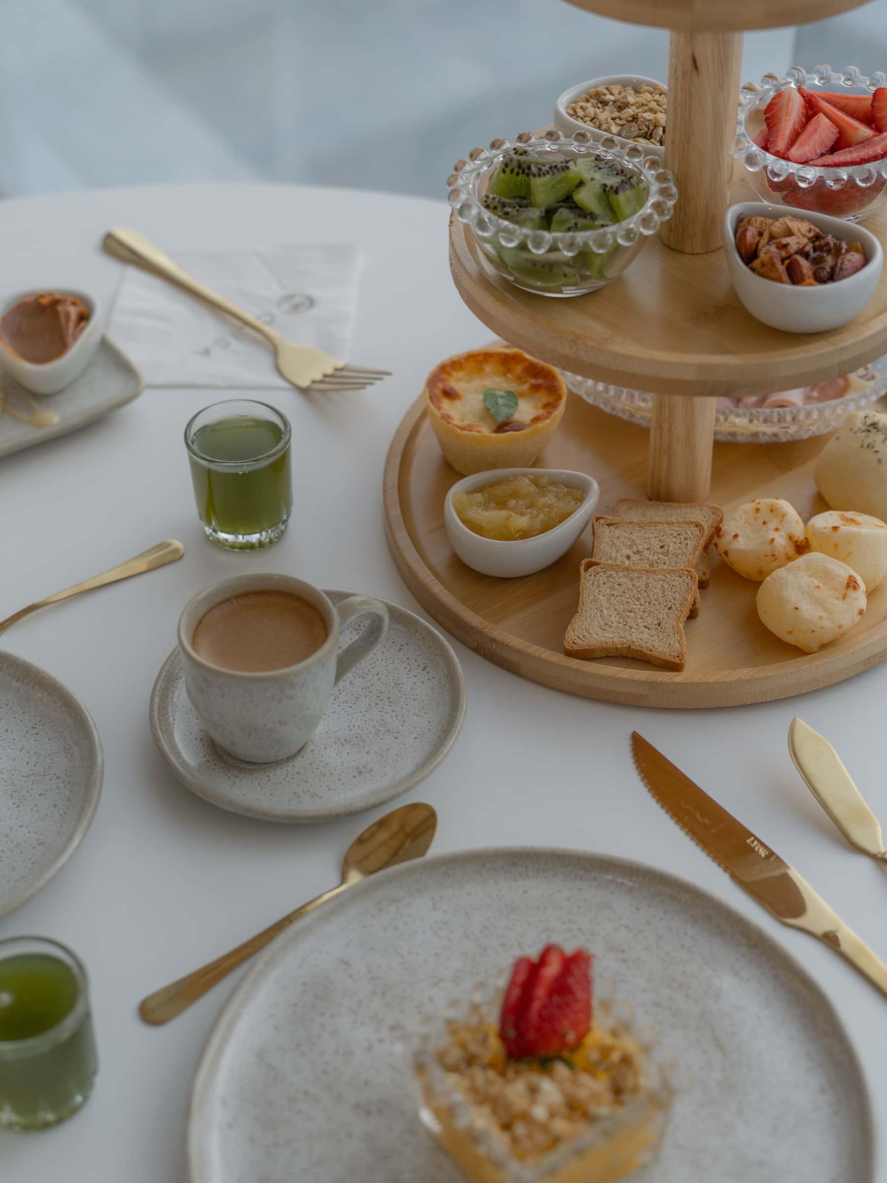 Mesa farta de café da manhã: torre de doces e frutas, xícara de café, suco verde e pratos de cerâmica clara