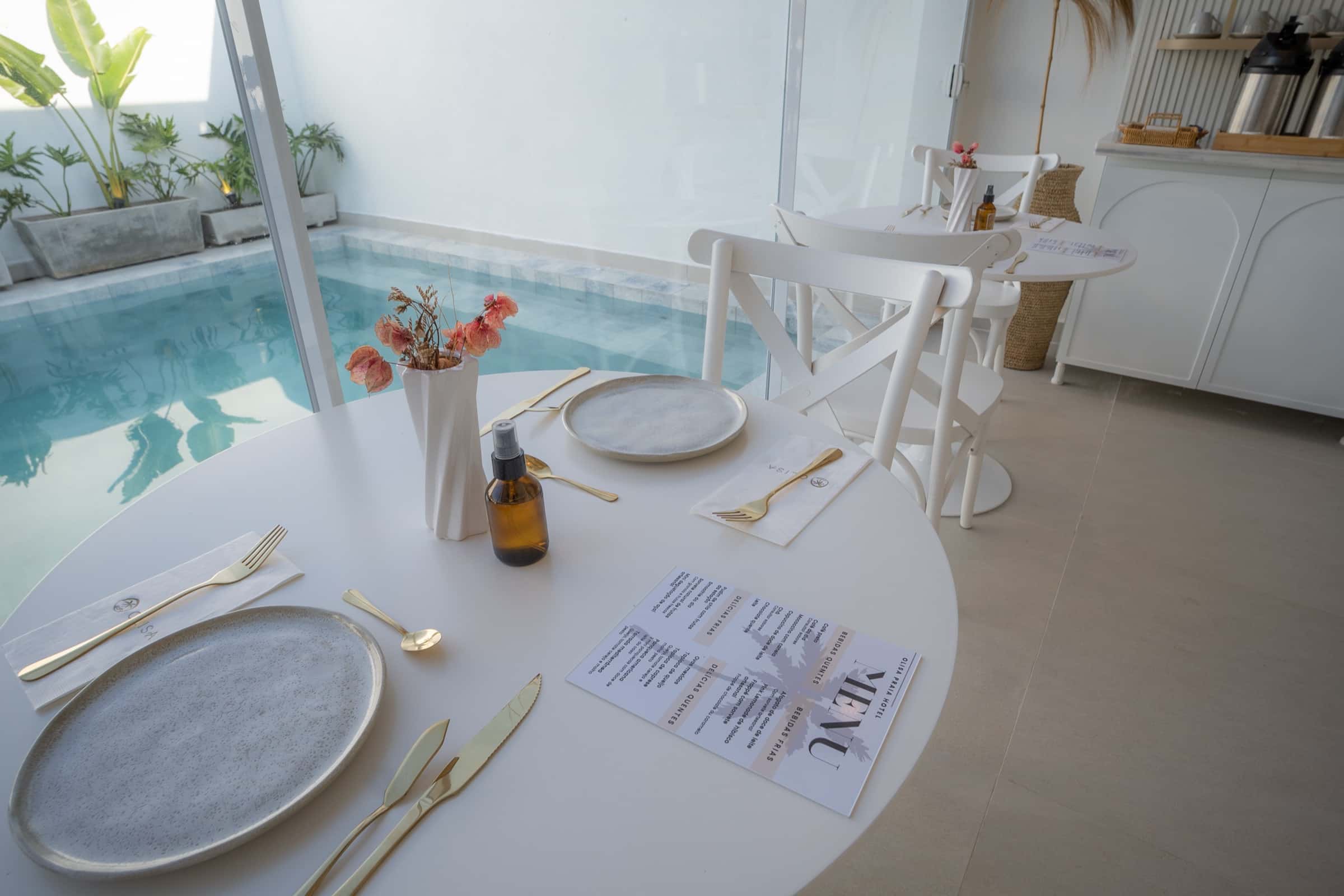 Mesa posta para o café da manhã à beira da piscina, com louça clara, talheres dourados e o cardápio do ALMA apoiado sobre a toalha