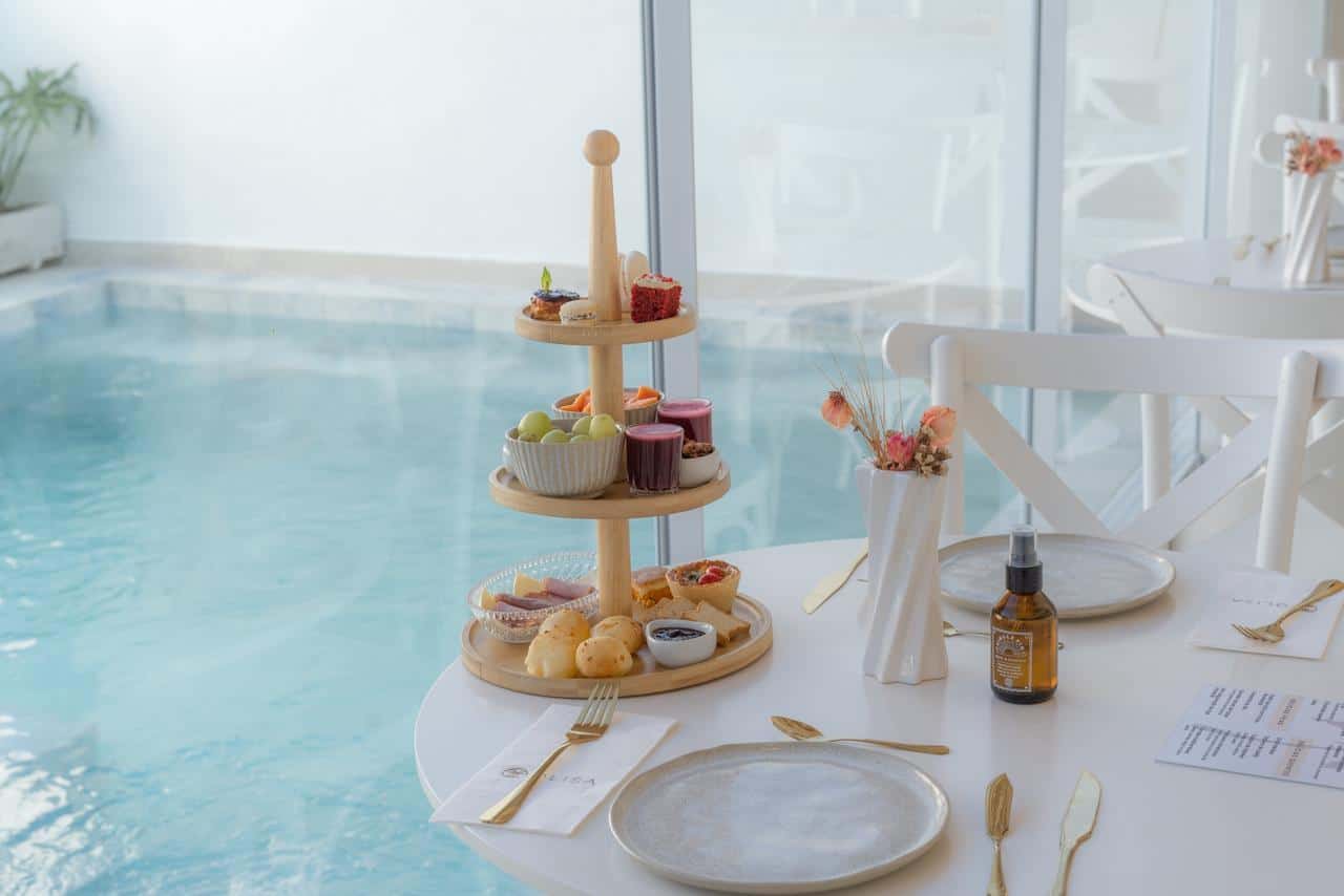 Torre de três andares com pão de queijo, tortinhas, frutas, iogurtes e doces sobre a mesa posta, com a água da piscina atrás do vidro do salão