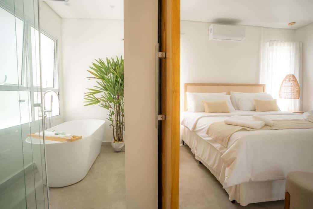 Banheira solta de borda alta ao lado de uma palmeira em vaso, com bandeja de madeira apoiada na borda, vista do quarto pela porta de vidro, e a cama king-size arrumada do outro lado