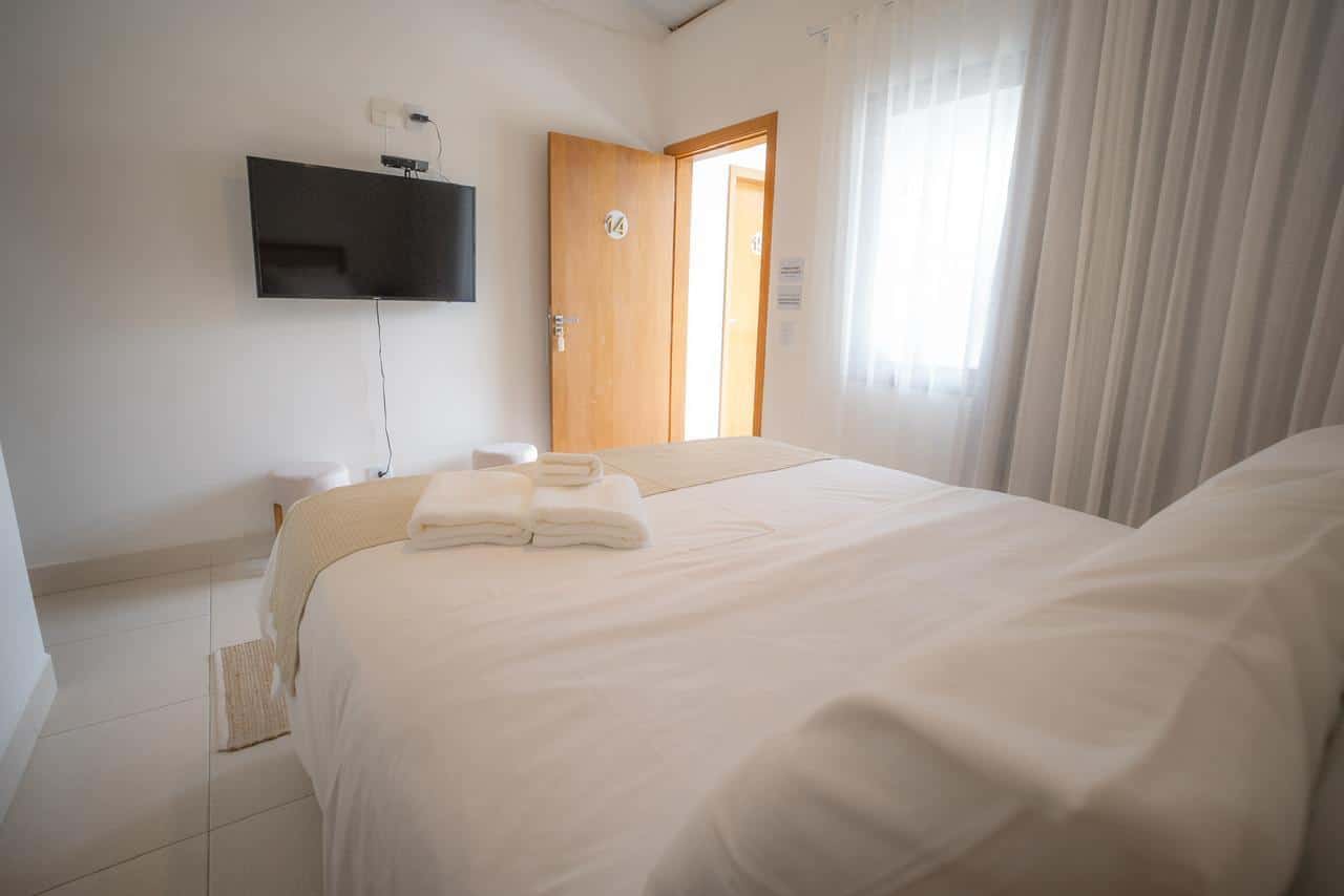 Quarto com televisão de tela plana na parede, porta de madeira aberta para o corredor, cortina de voil na janela e toalhas dobradas sobre a cama