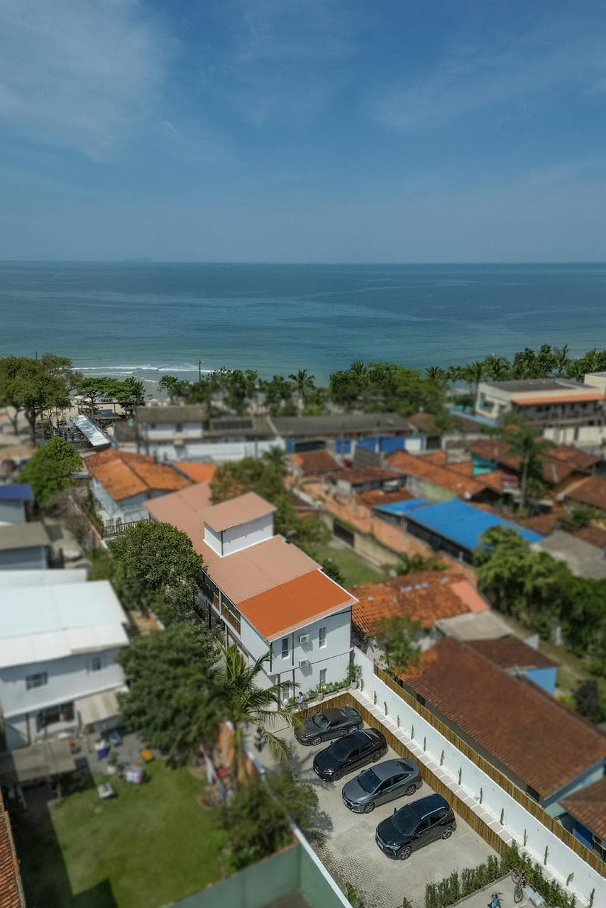 Vista aérea do bairro: o telhado de duas águas do hotel e o pátio de estacionamento em primeiro plano, os telhados de Maresias em volta, a faixa de areia e o mar aberto ocupando o fundo do quadro