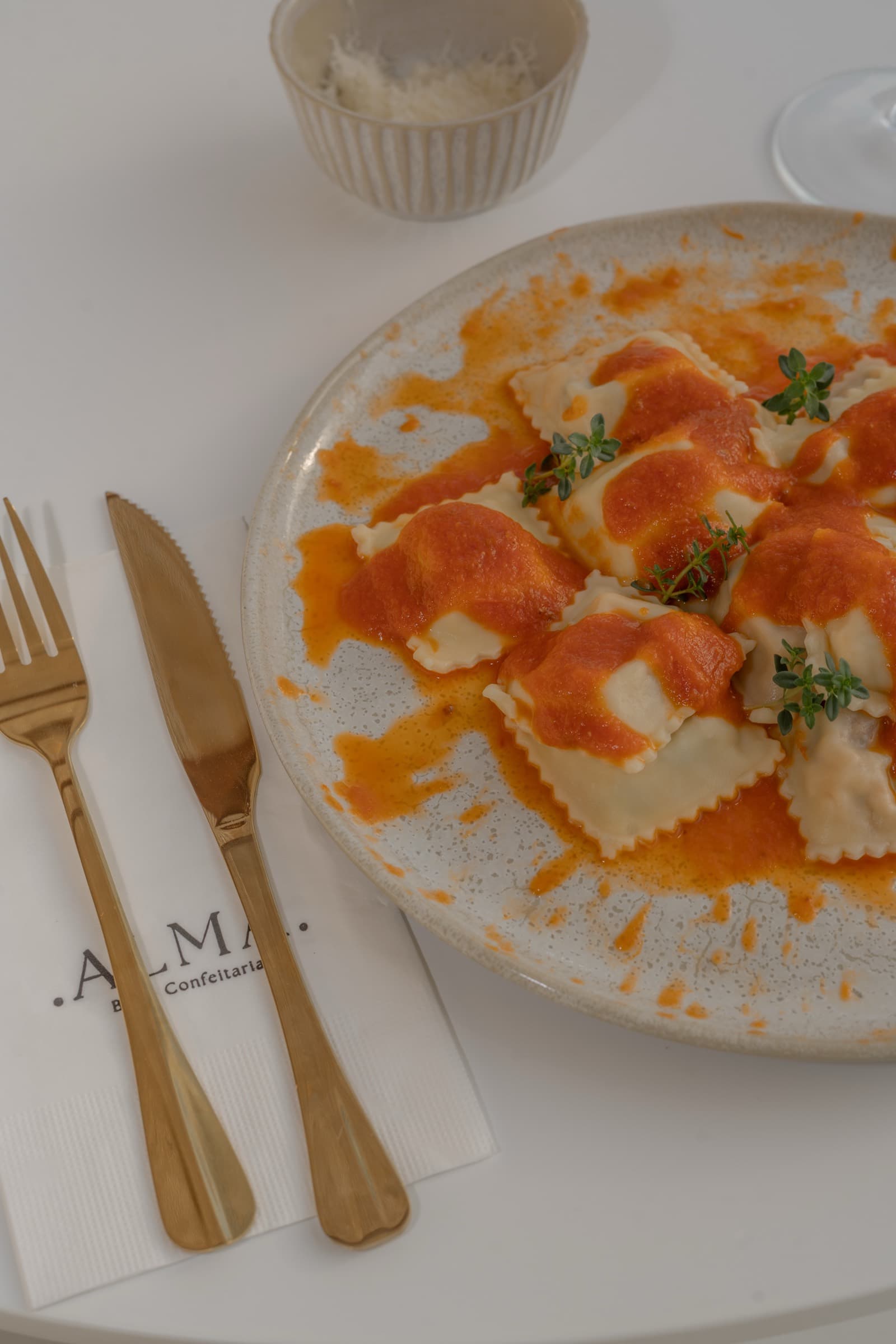 Prato de ravioli ao molho vermelho sobre cerâmica clara, com talheres dourados e um guardanapo de linho com o logotipo ALMA bordado