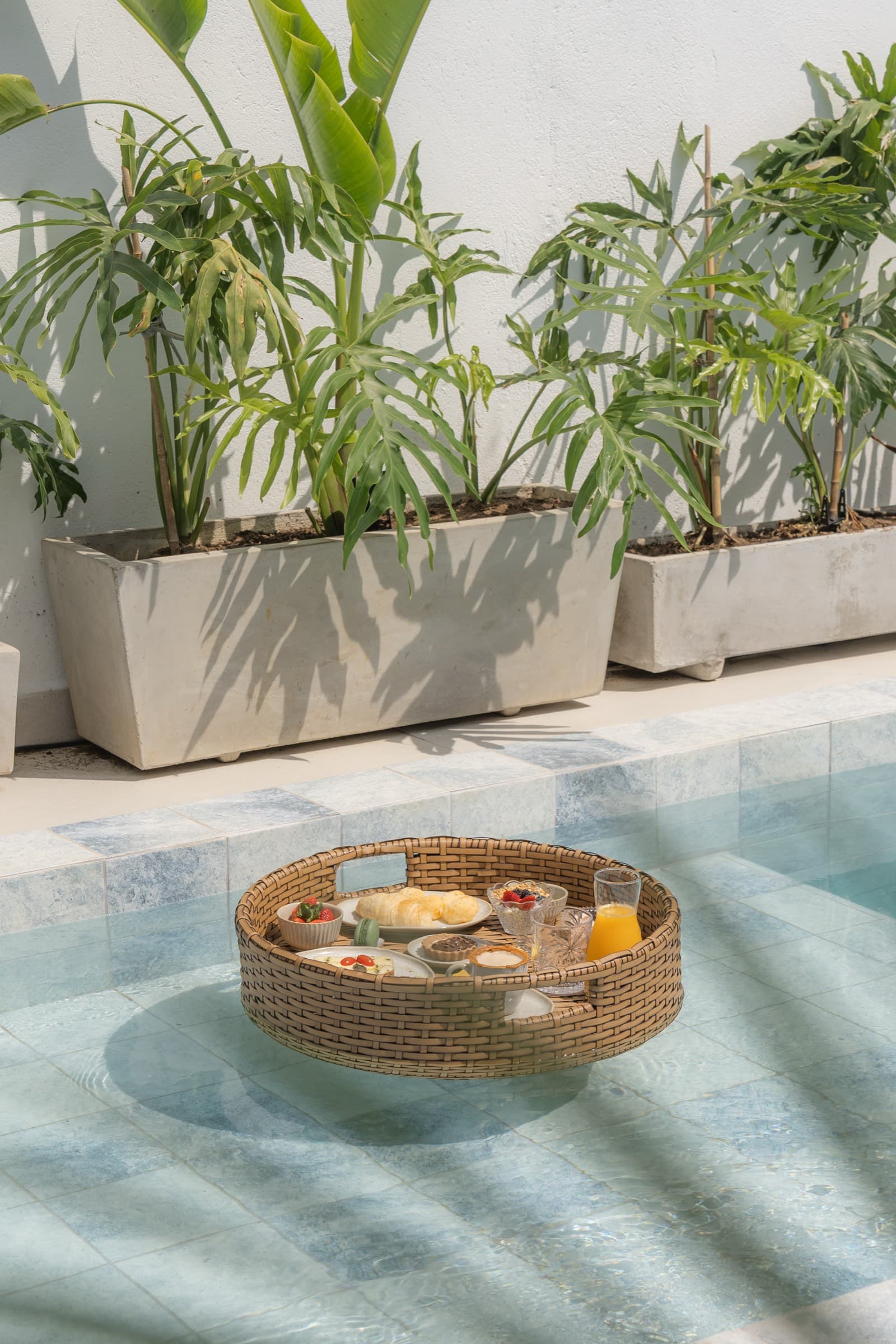 Bandeja de vime com café da manhã flutuando na água rasa da piscina, com vasos de folhagem tropical e a sombra das folhas no muro caiado