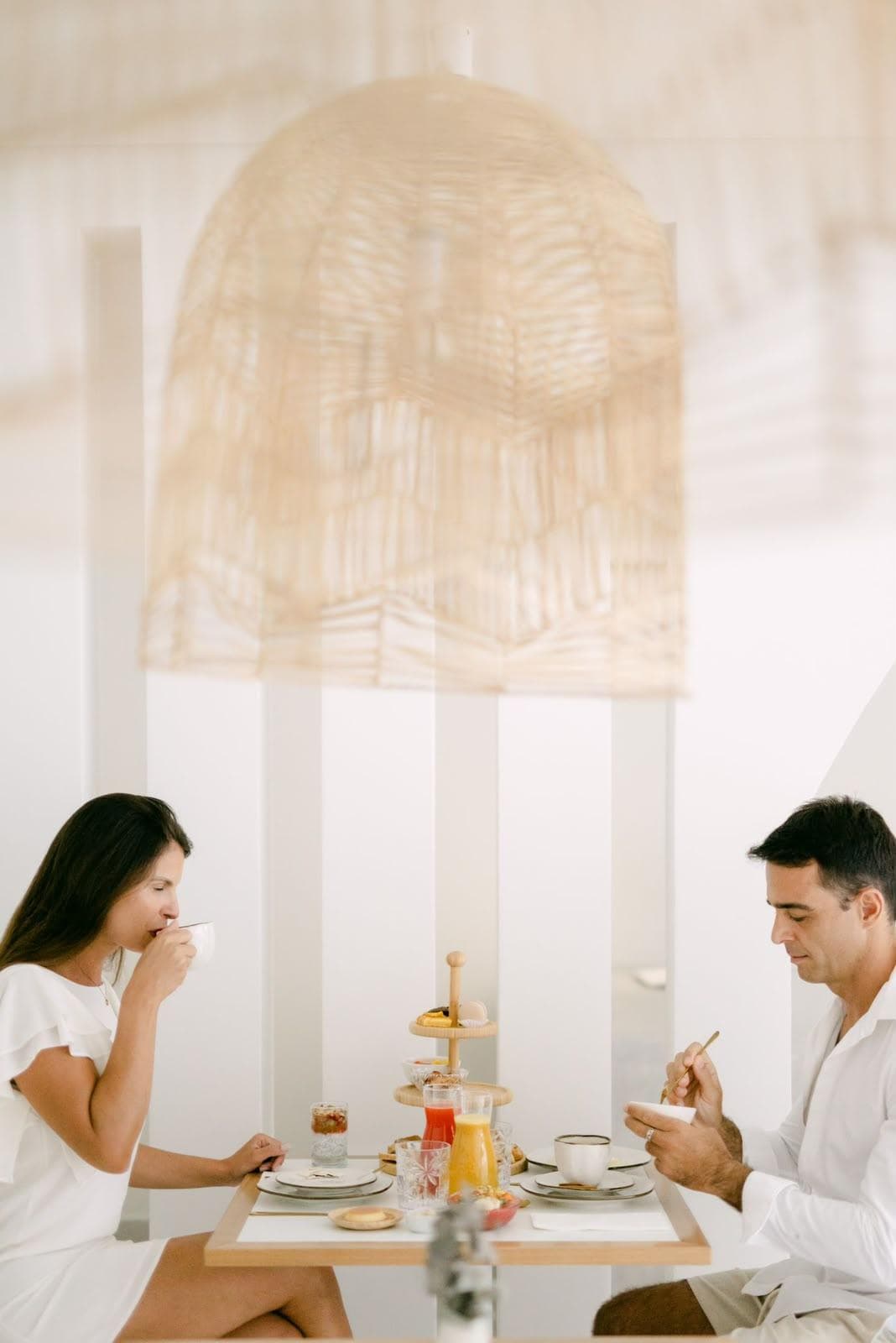 Casal à mesa do café da manhã no salão branco: ela toma café na xícara, ele se serve de uma tigela, com a torre de três andares, os sucos e a louça entre os dois e um grande pendente de fibra trançada por cima