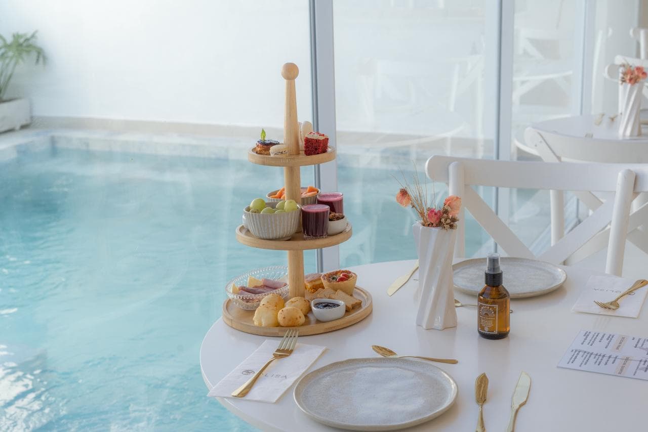 Torre de três andares com pão de queijo, tortinhas, frutas, iogurtes e doces sobre a mesa posta, com a água da piscina atrás do vidro do salão