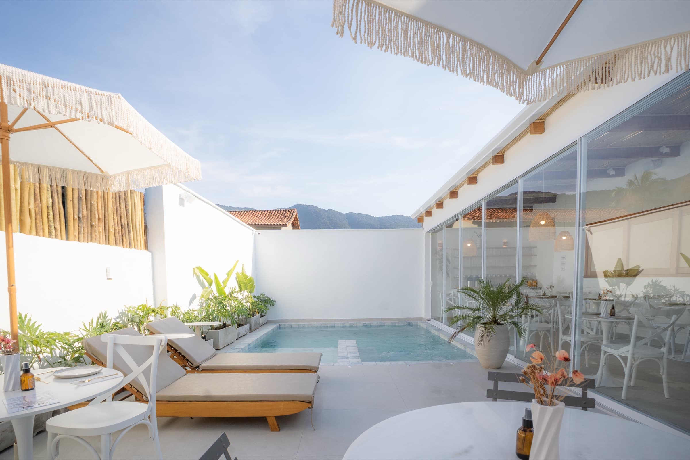Espreguiçadeiras de madeira e guarda-sóis de franja à beira da piscina, com o muro branco, a serra e o céu aberto ao fundo