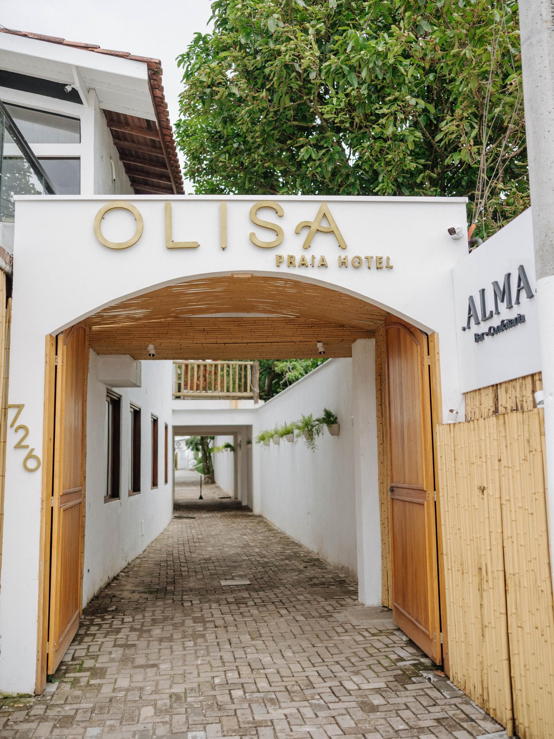 Portal de entrada do hotel: arco caiado com as letras douradas OLISA PRAIA HOTEL e o número 726, forro de bambu, portas de madeira abertas para o caminho de pedra e a placa do ALMA Bar + Confeitaria ao lado