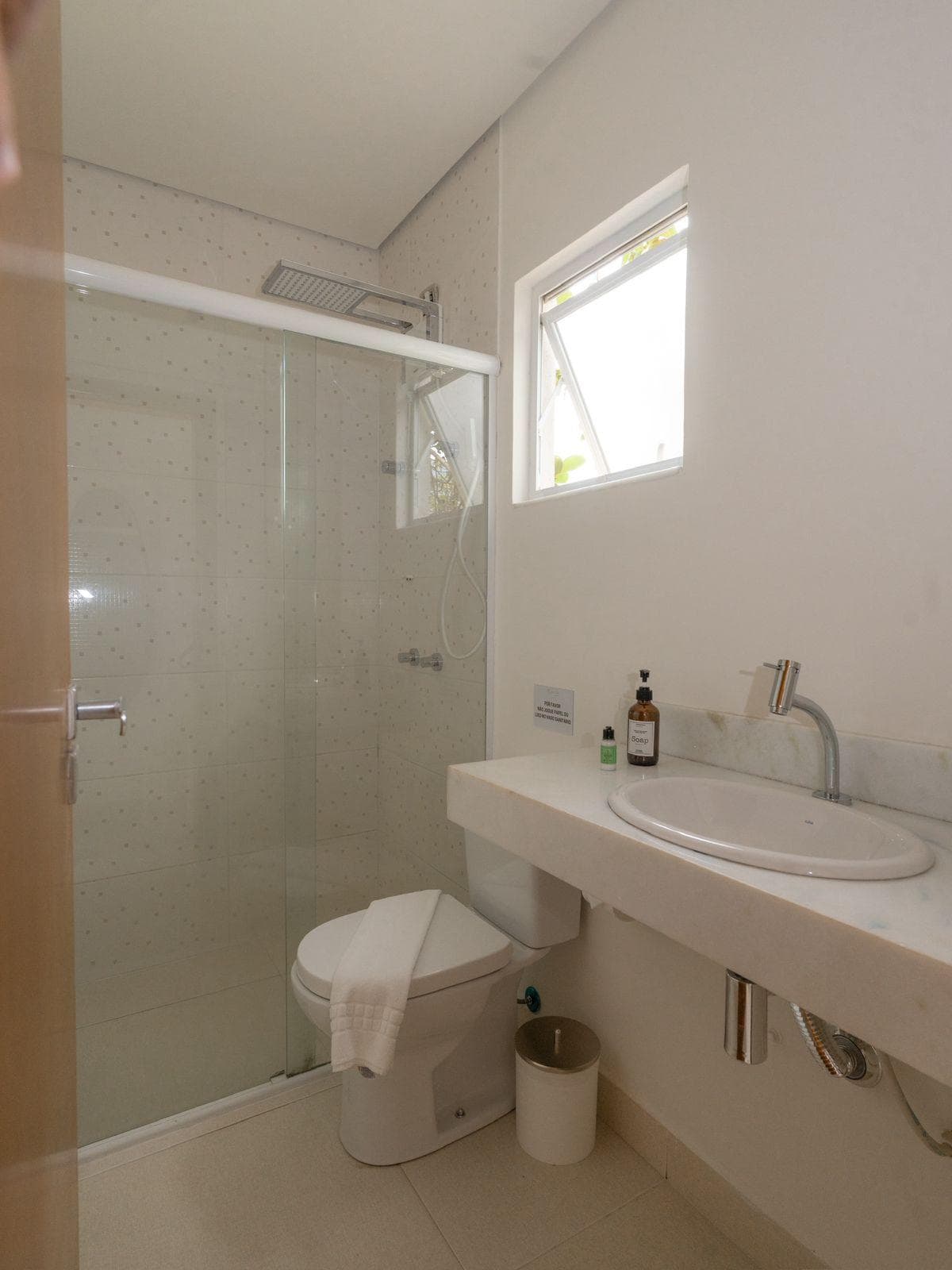 Banheiro da suíte: box de vidro com ducha de teto, vaso sanitário, bancada de mármore com cuba de louça, sabonete líquido e janela basculante aberta