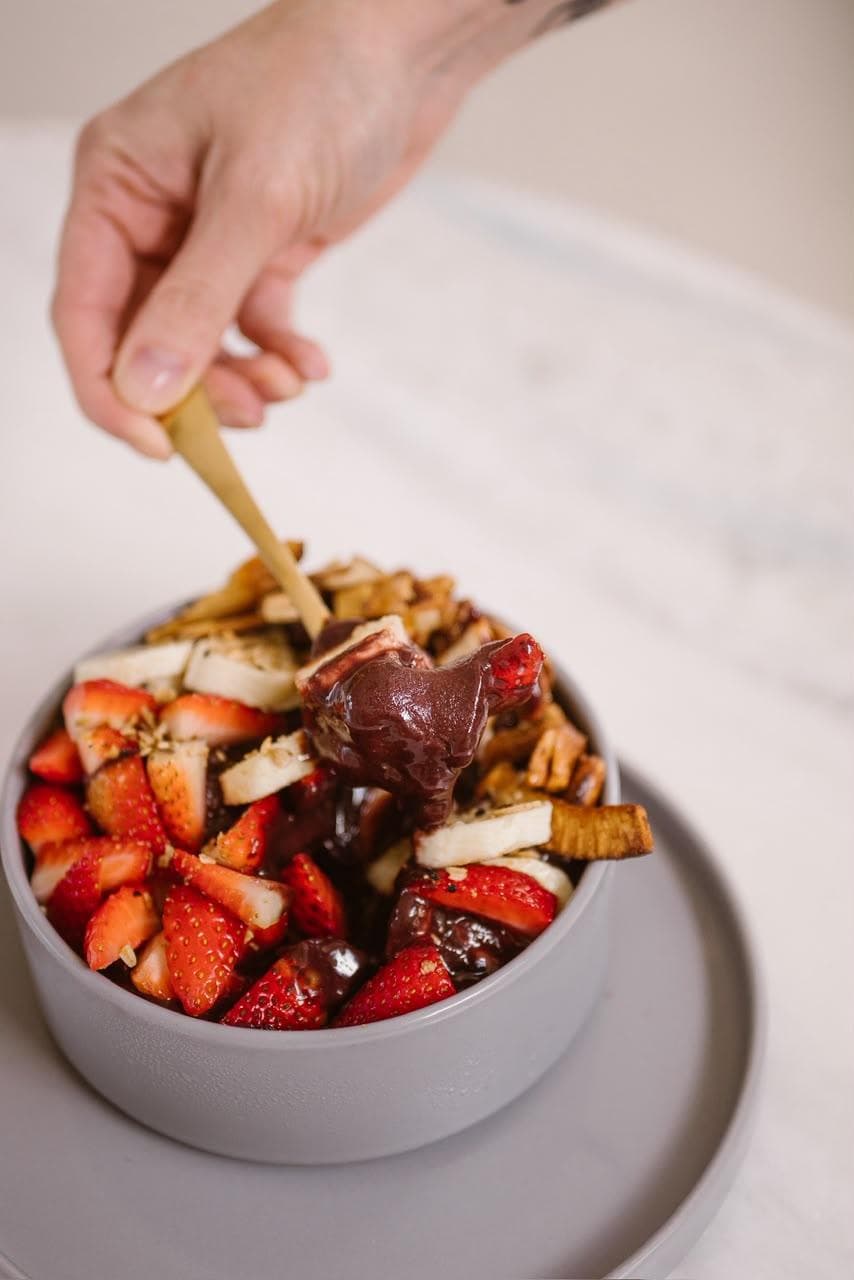 Tigela cinza de açaí com morangos, banana e granola, com uma colher de madeira recolhendo a polpa