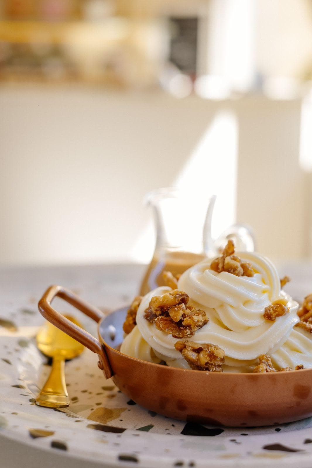 Chantilly em ondas numa panelinha de cobre, coberto de nozes caramelizadas, com uma jarrinha de doce de leite atrás e um prato de terrazzo por baixo
