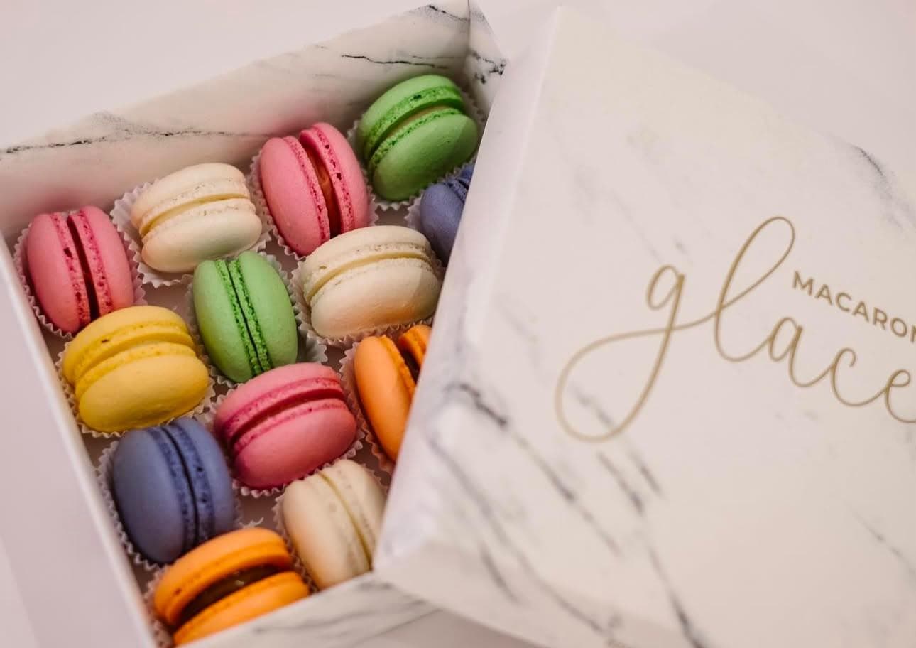 Caixa aberta de macarons coloridos — rosa, verde, amarelo, lilás, laranja e branco — em forminhas de papel, com a tampa de estampa de mármore trazendo a marca Macarons Glace em letras douradas