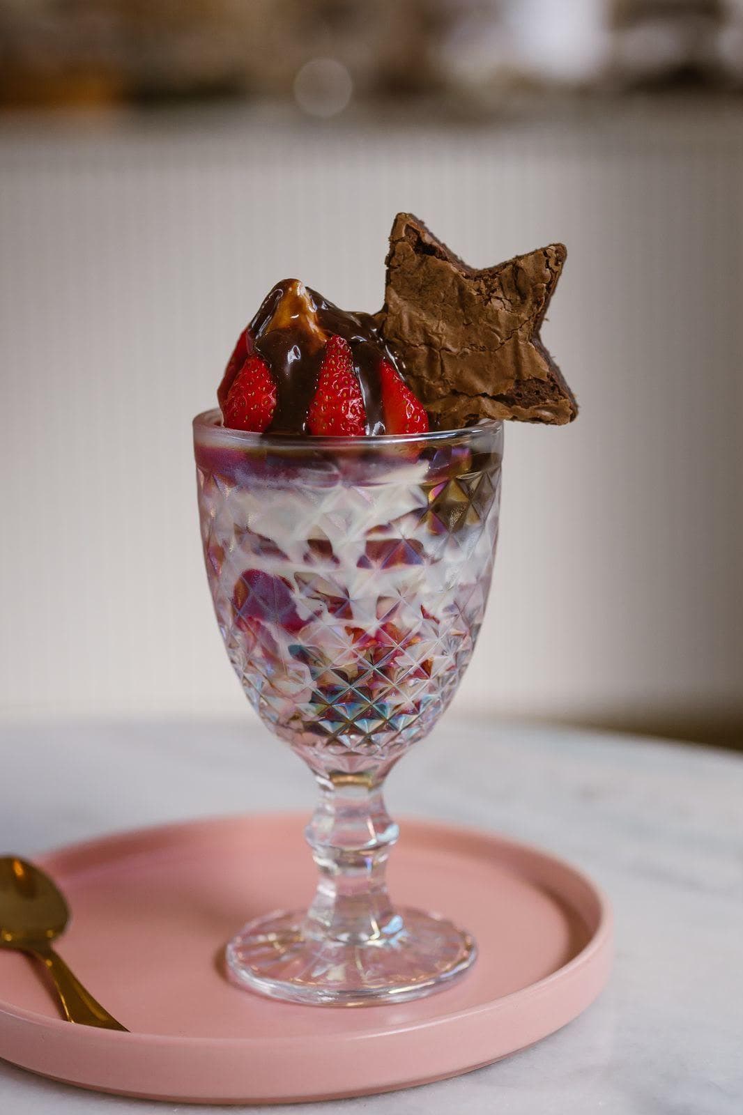 Taça de cristal lapidado com creme e morangos em camadas, coberta por calda de chocolate e com um pedaço de brownie apoiado na borda, sobre prato rosa