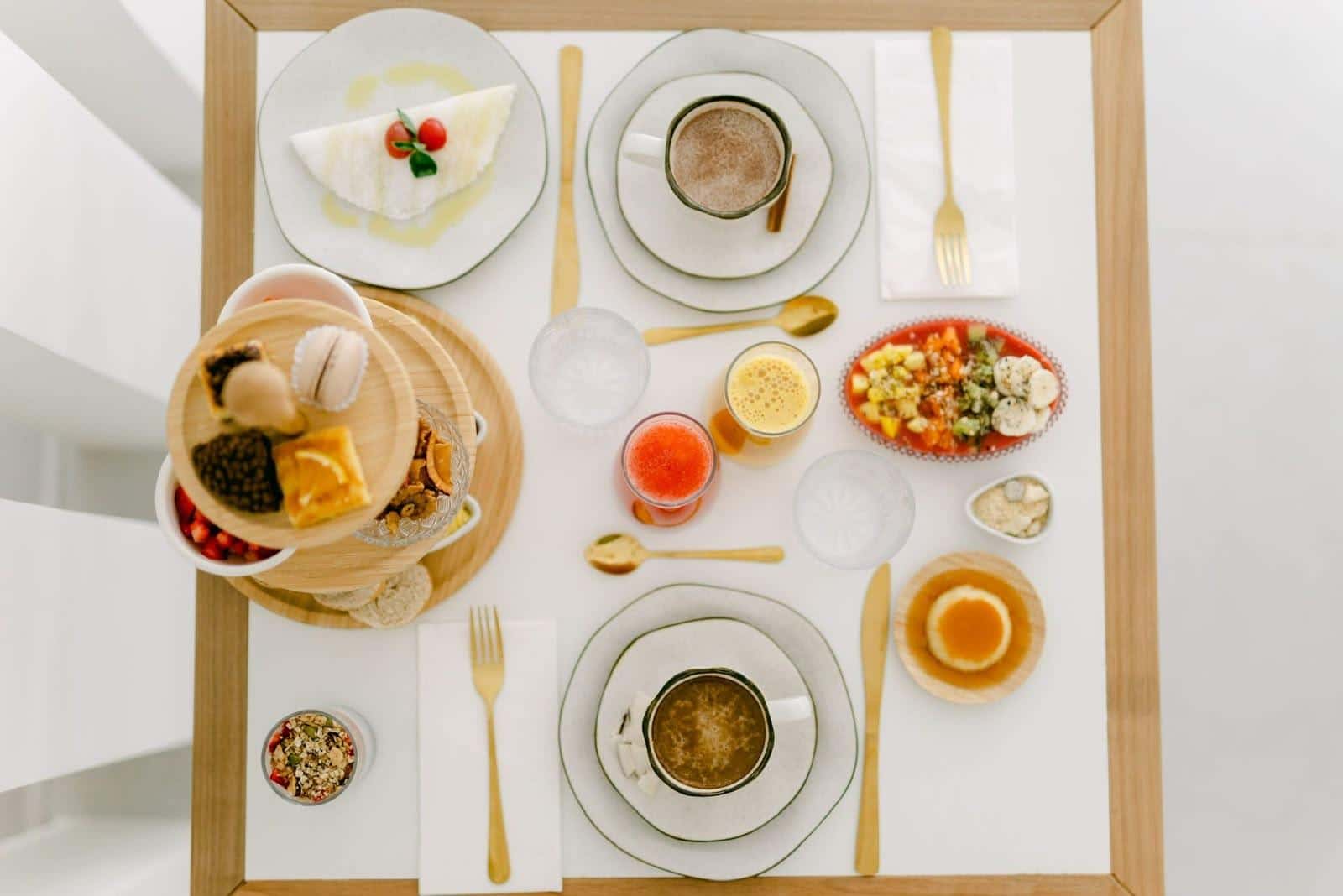 Mesa quadrada do café da manhã vista exatamente de cima: tapioca com tomatinhos, dois cafés, sucos de laranja e de melancia, travessa de frutas com granola, pudim de caramelo e a torre de doces ocupando o canto