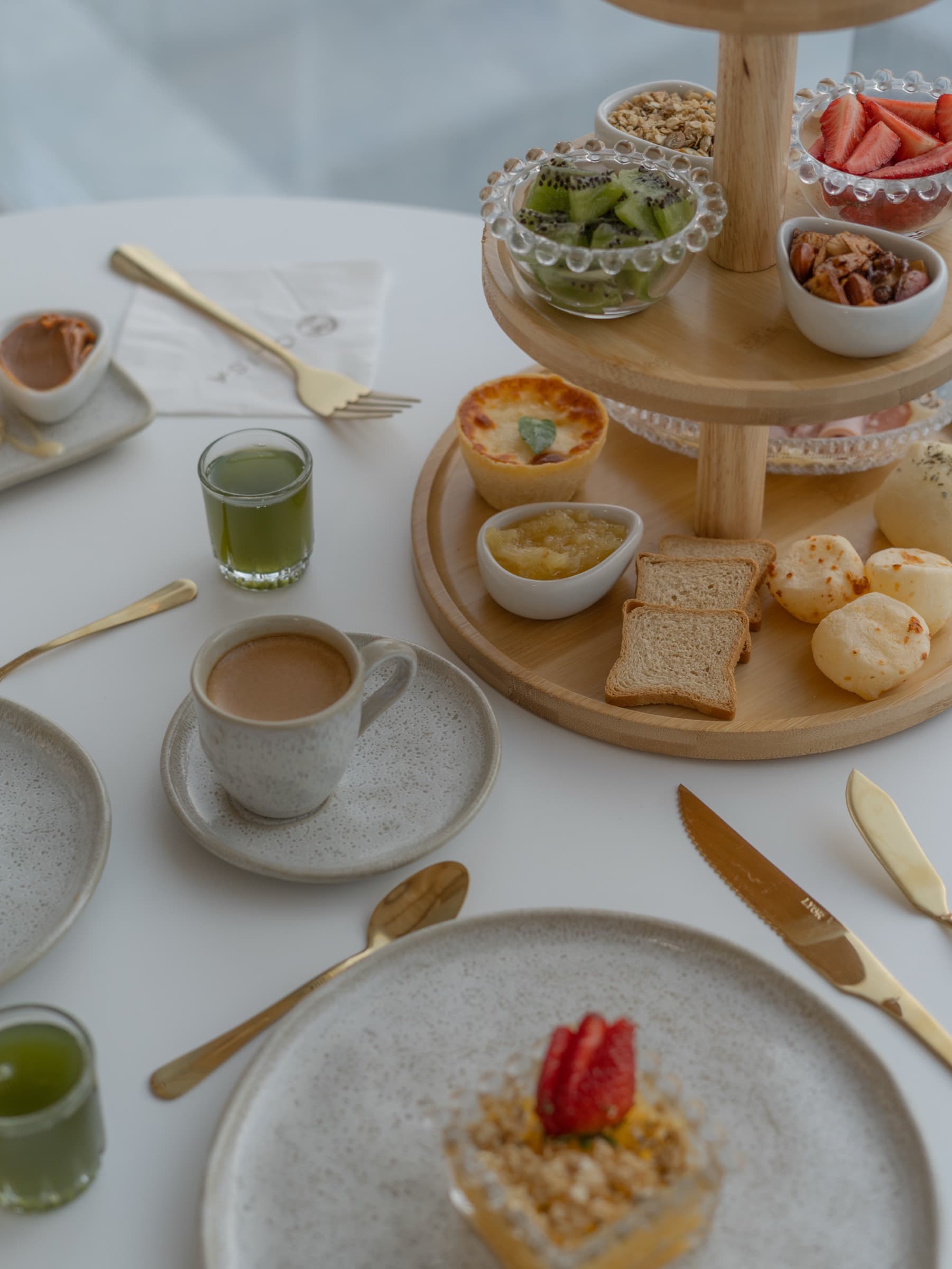 Mesa farta de café da manhã: torre de doces e frutas, xícara de café, suco verde e pratos de cerâmica clara