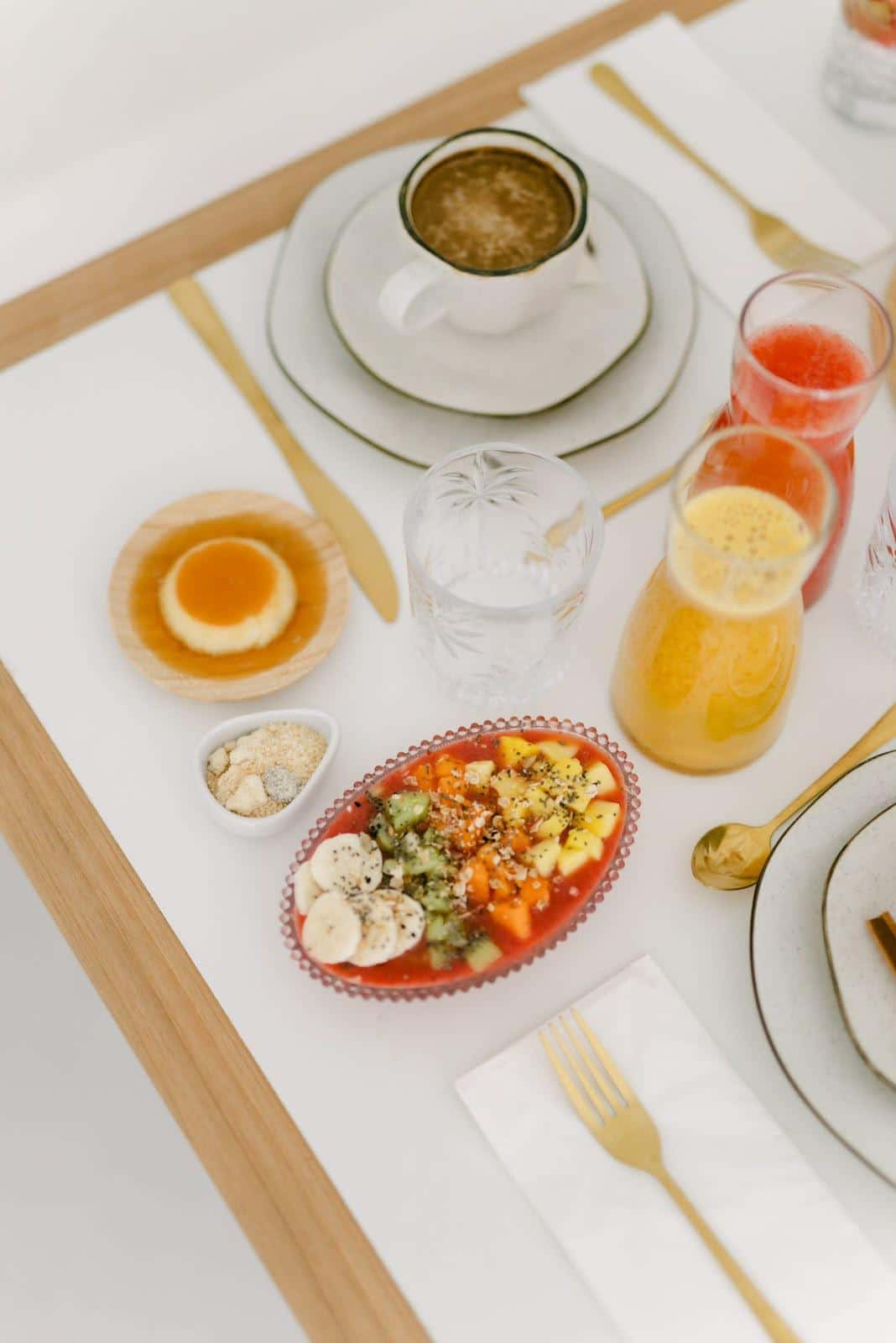 Mesa posta do café da manhã vista do alto: xícara de café, pudim de caramelo numa tigelinha de madeira, travessa de frutas com granola e chia e duas jarras de suco de laranja e de melancia
