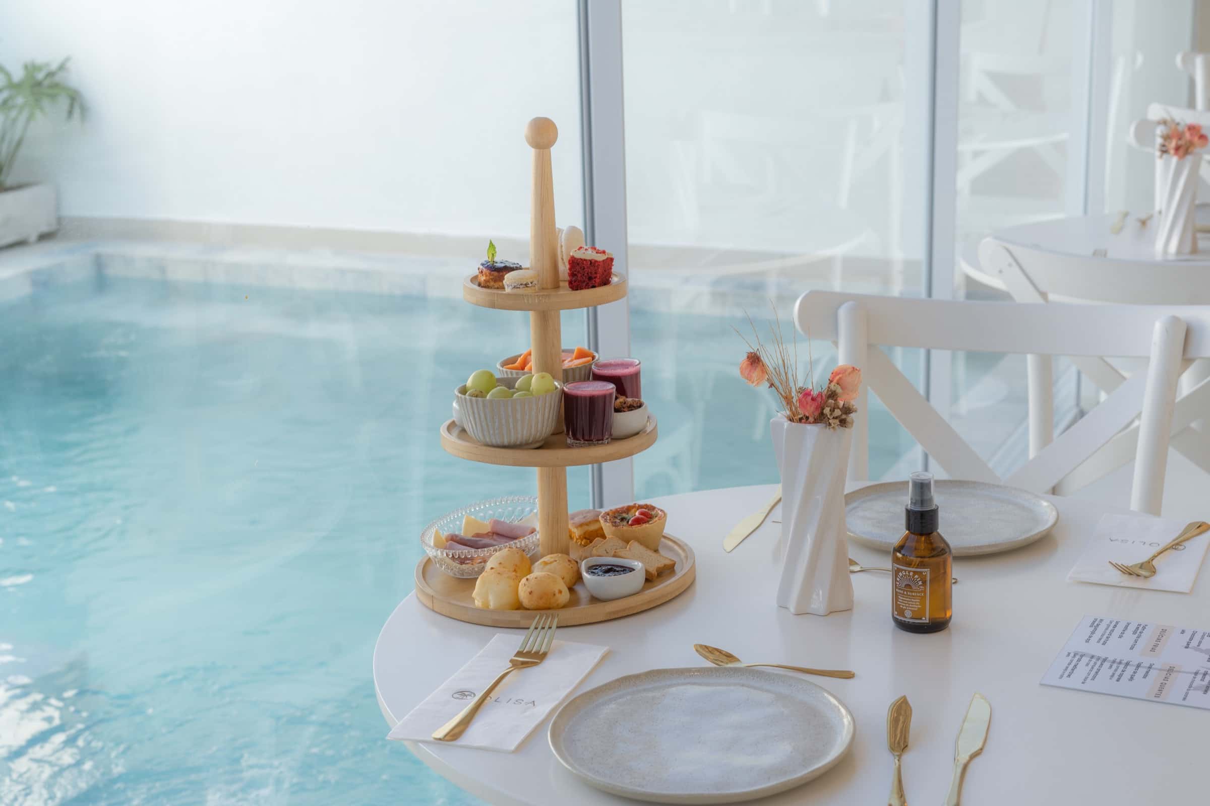 Torre de três andares com doces, pães e frutas montada na mesa, com a água da piscina logo atrás