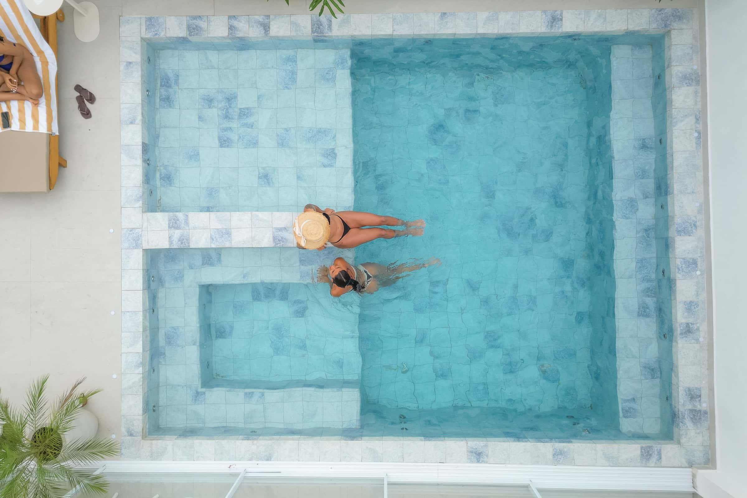 Vista aérea da piscina em L, revestida de azulejo azul artesanal, com duas mulheres de biquíni na lâmina rasa
