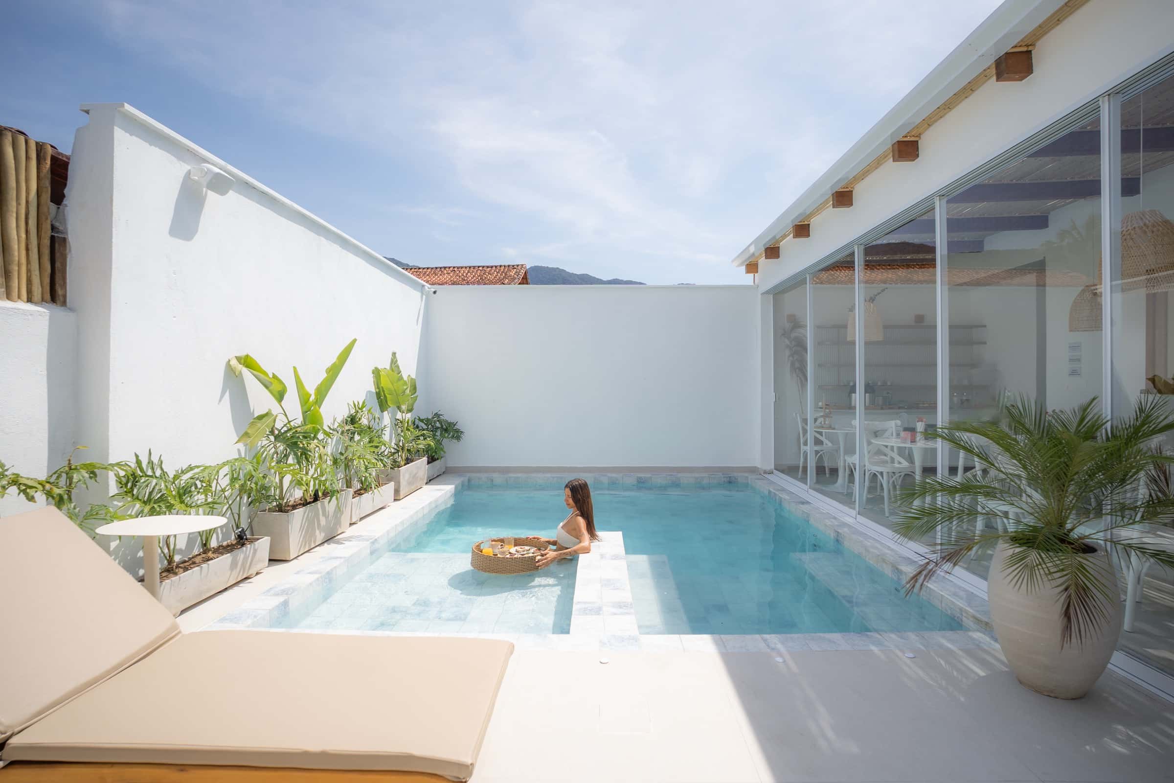 Pátio da piscina entre muros brancos caiados: uma hóspede na água ao lado de uma bandeja flutuante de vime, o salão envidraçado à direita e a serra atrás do telhado