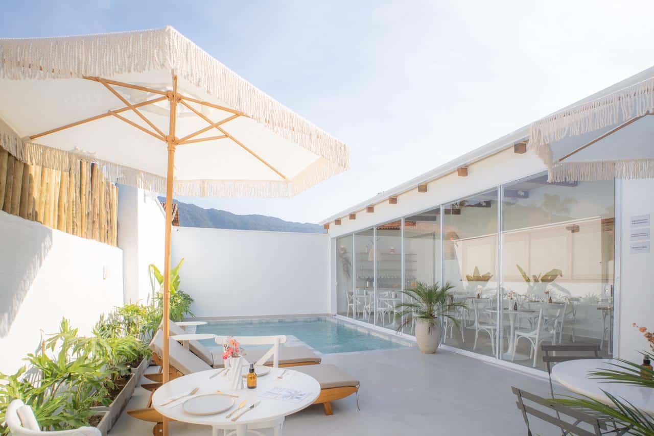 Pátio da piscina contra a luz: guarda-sol de franja sobre a mesa posta, espreguiçadeiras de madeira, a serra por cima do muro caiado e o salão envidraçado ocupando o lado direito