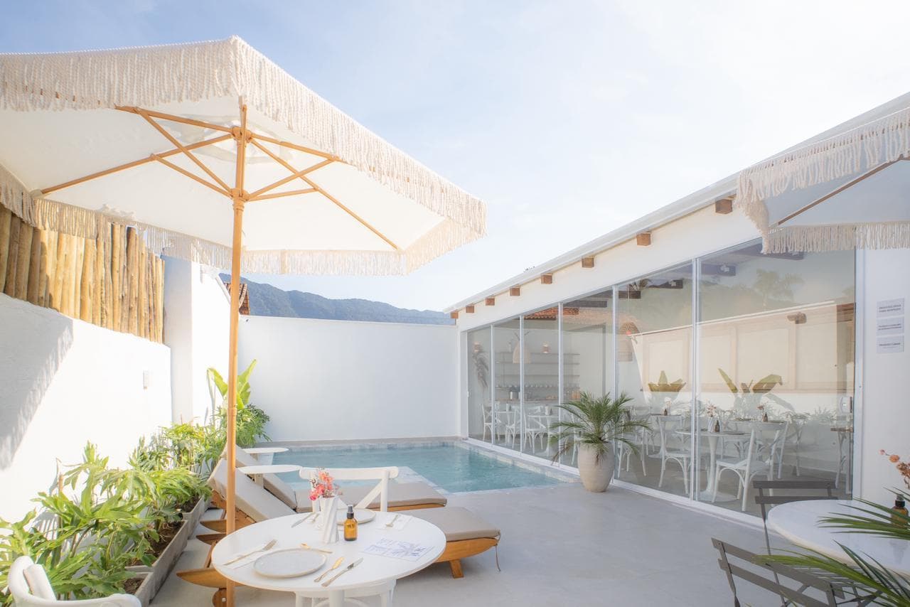 Pátio da piscina contra a luz: guarda-sol de franja sobre a mesa posta, espreguiçadeiras de madeira, a serra por cima do muro caiado e o salão envidraçado ocupando o lado direito