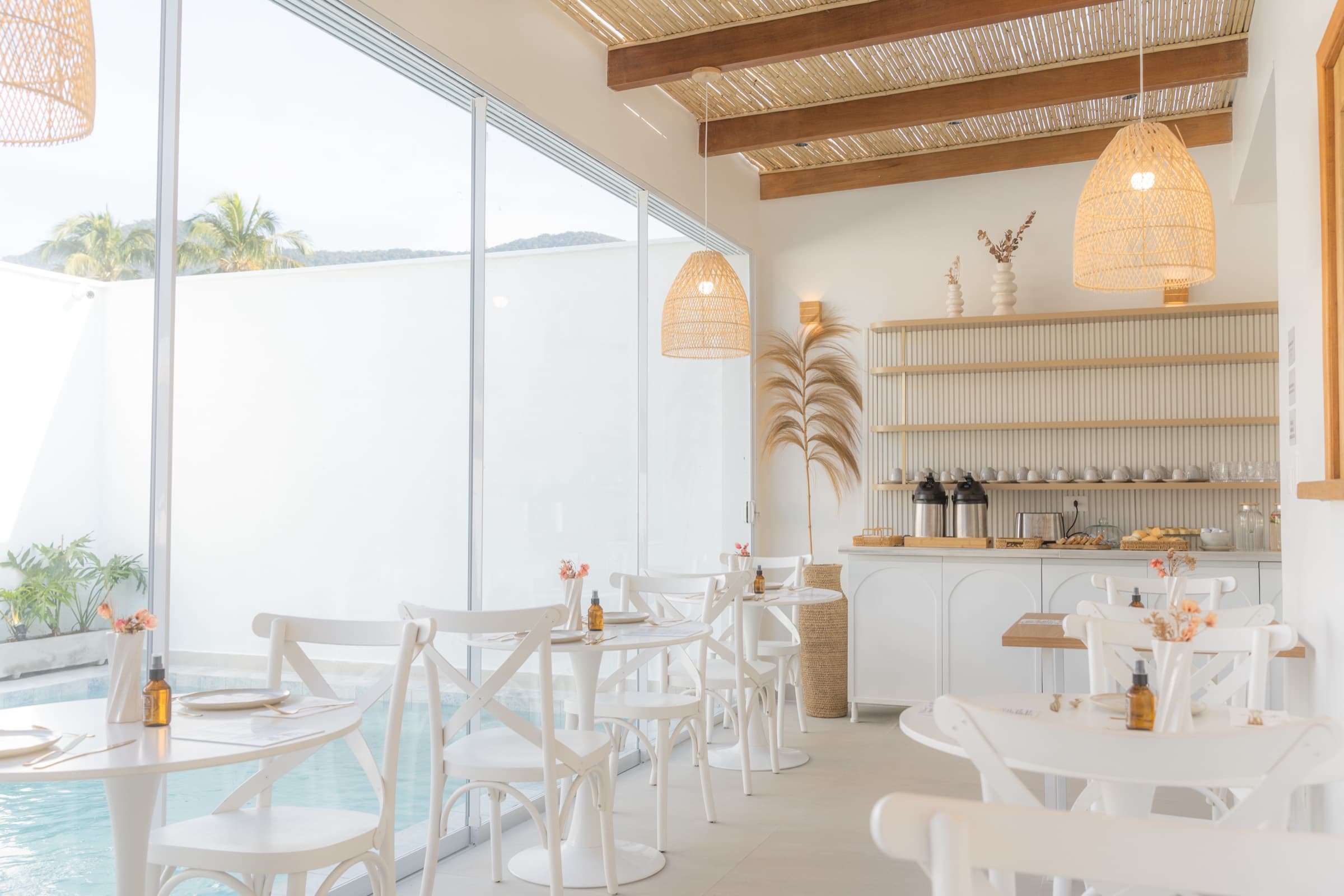 Salão branco com vidro do chão ao teto para a piscina, mesas redondas, cadeiras bistrô brancas, pendentes de fibra trançada e viga de madeira aparente