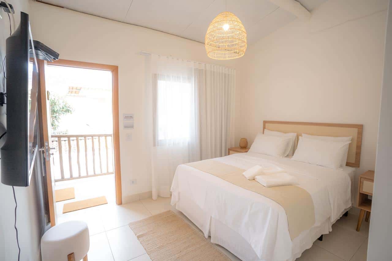 Quarto com a porta da varanda aberta para o guarda-corpo de madeira roliça, pendente de fibra trançada sobre a cama de casal e televisão na parede oposta